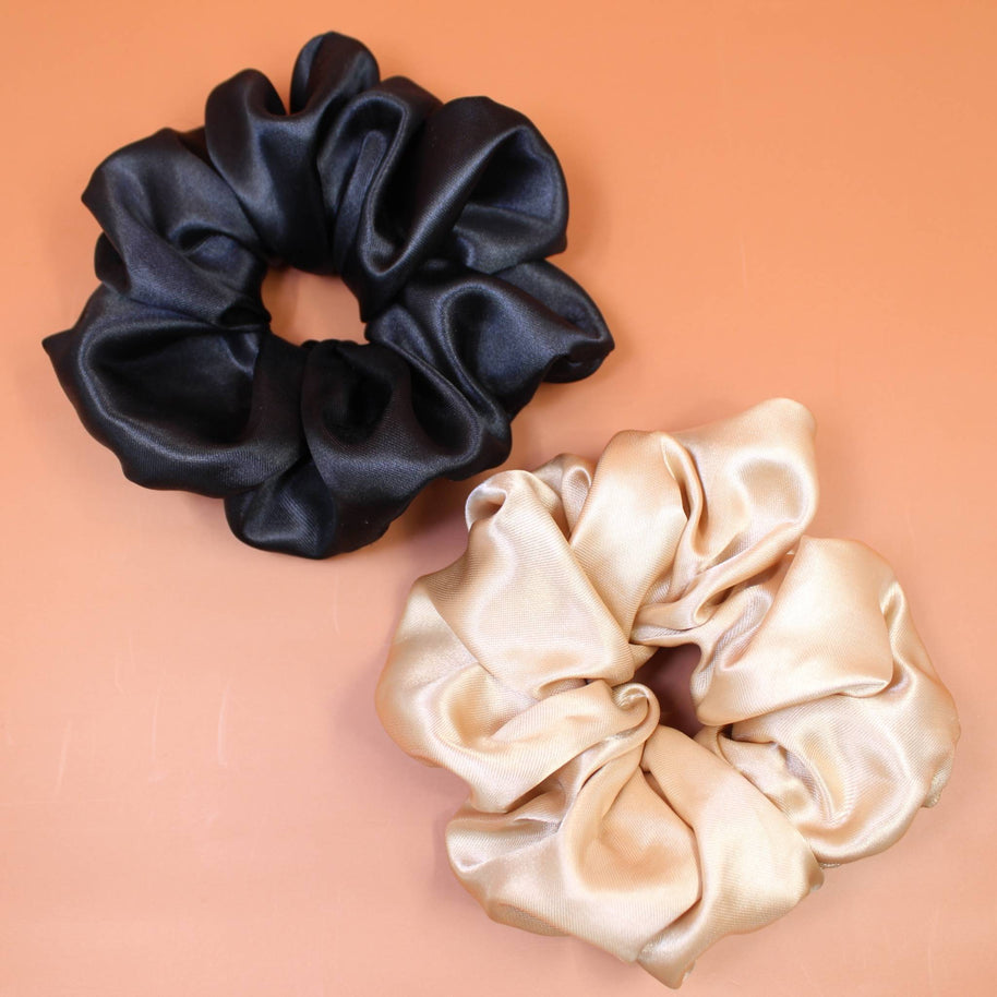 Afro Scrunchie Satin Beige und Afro Scrunchie Satin Schwarz, beide liegend nebeneinander auf einen braunen Hintergrund