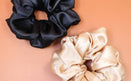 Afro Scrunchie Satin Beige und Afro Scrunchie Satin Schwarz, beide liegend nebeneinander auf einen braunen Hintergrund
