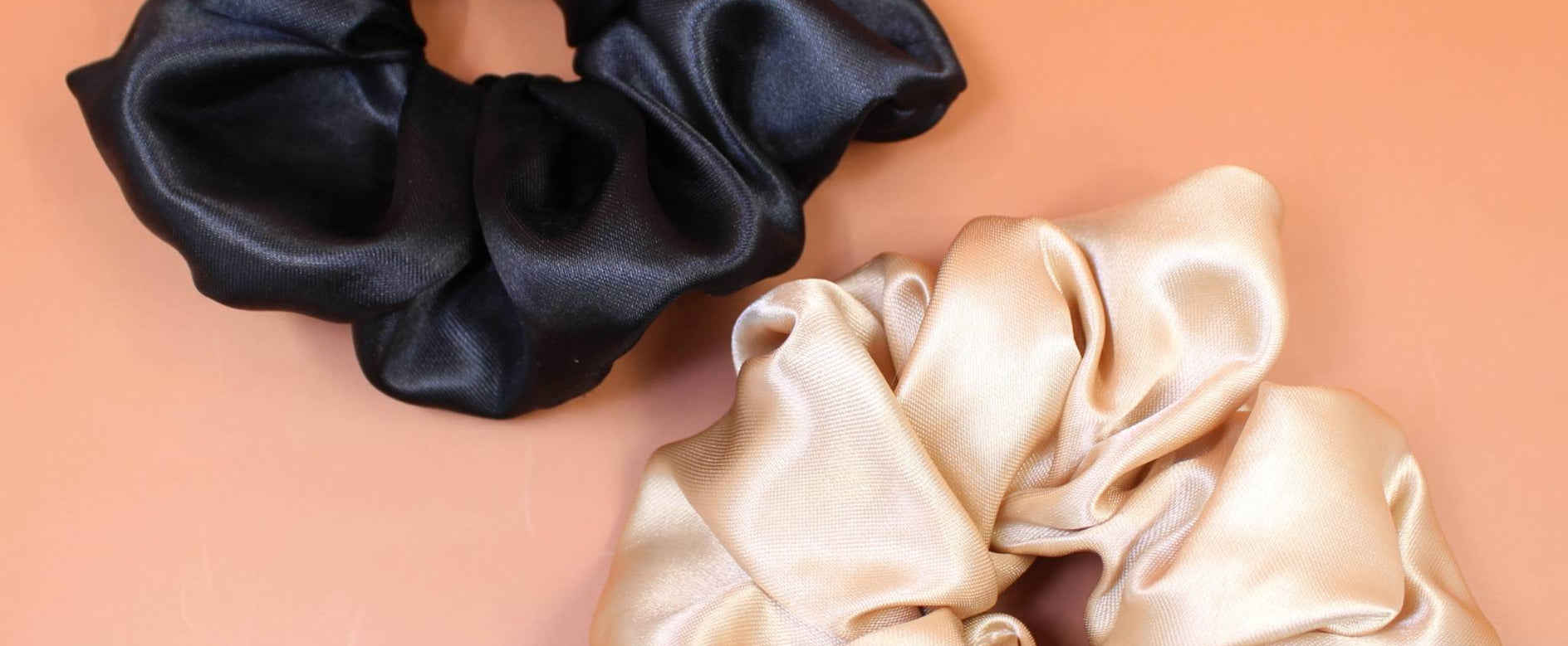 Afro Scrunchie Satin Beige und Afro Scrunchie Satin Schwarz, beide liegend nebeneinander auf einen braunen Hintergrund