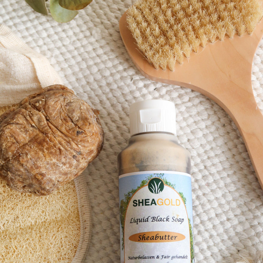 Das Hautpflegeprodukt SHEA GOLD Black Soap 100g und  SHEA GOLD Liquid Black Soap 240ml  in der Vorderansicht auf einem beigen Untergrund mit einer Bürste und einem Schwamm im Hintergrund.