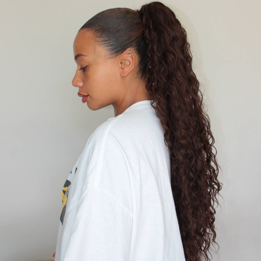 Junge Frau trägt den DREAM HAIR Ponytail Diborra Curly in der Seitenansicht vor einem hell-grauen Hintergrund.