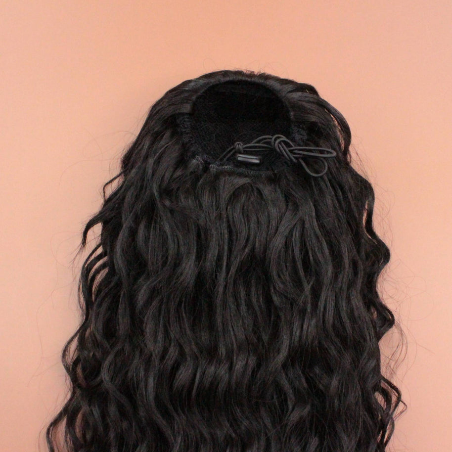 DREAM HAIR Ponytail Diborra Curly, Farbe 2 (dunkelbraun) in der Rückansicht vor einem braun-orangen Hintergrund.