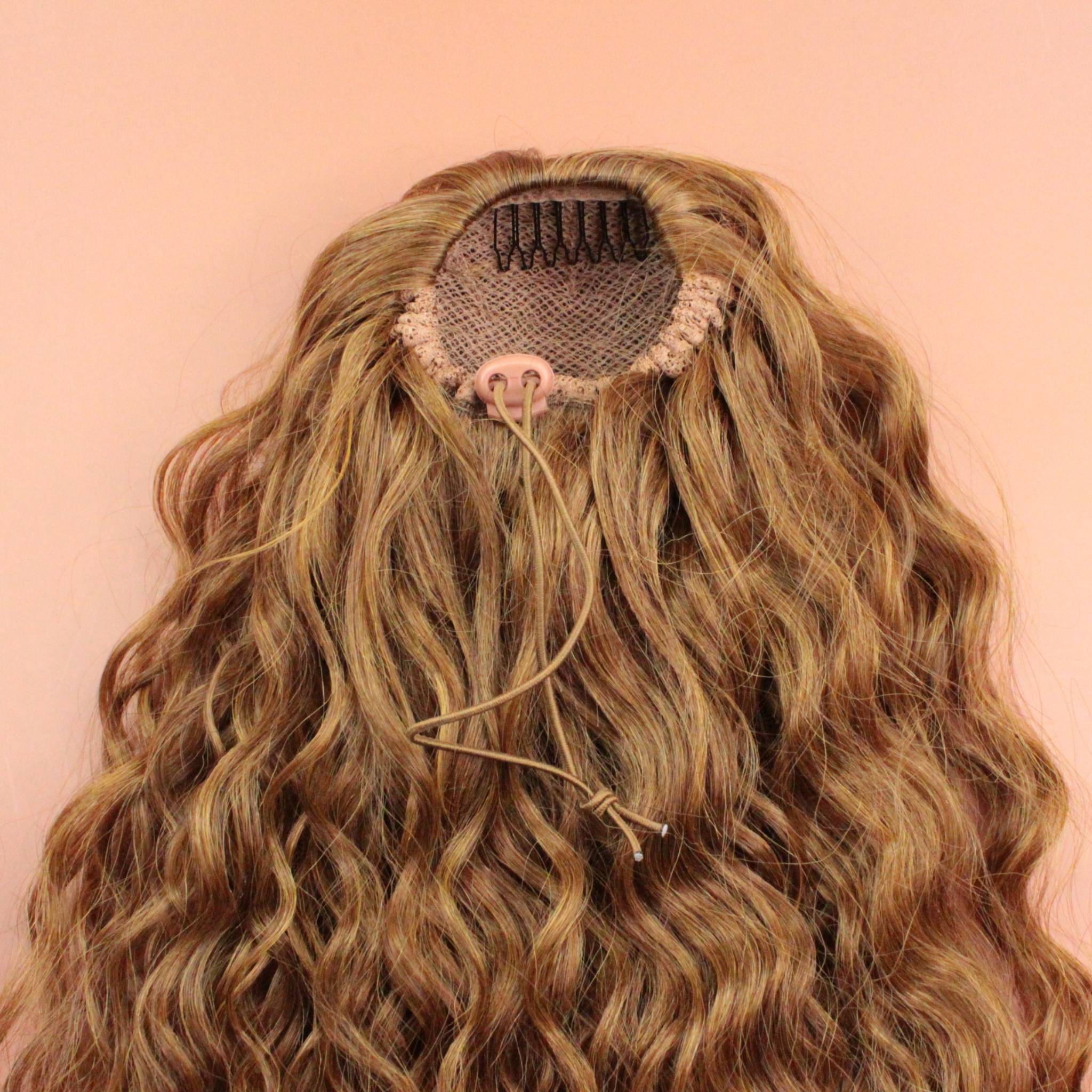 DREAM HAIR Ponytail Diborra Curly, Farbe 27 (goldblond) in der Rückansicht vor einem braun-orangen Hintergrund.