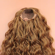 DREAM HAIR Ponytail Diborra Curly, Farbe 27 (goldblond) in der Rückansicht vor einem braun-orangen Hintergrund.