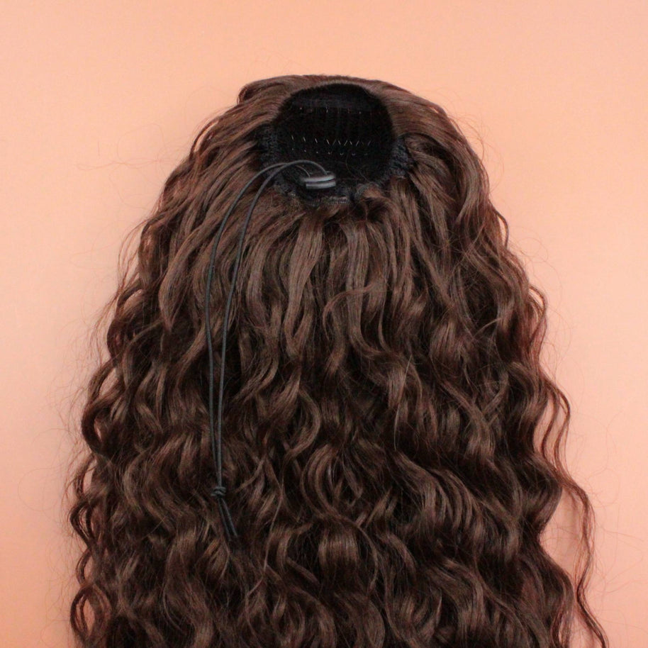 DREAM HAIR Ponytail Diborra Curly, Farbe 6 (mittelbraun) in der Rückansicht vor einem braun-orangen Hintergrund.