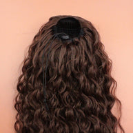 DREAM HAIR Ponytail Diborra Curly, Farbe 6 (mittelbraun) in der Rückansicht vor einem braun-orangen Hintergrund.