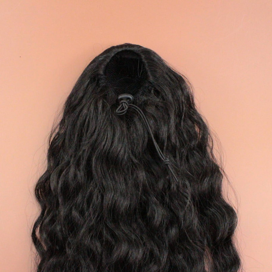 DREAM HAIR Ponytail Diborra Curly, Farbe 1B (schwarz) in der Rückansicht vor einem braun-orangen Hintergrund.