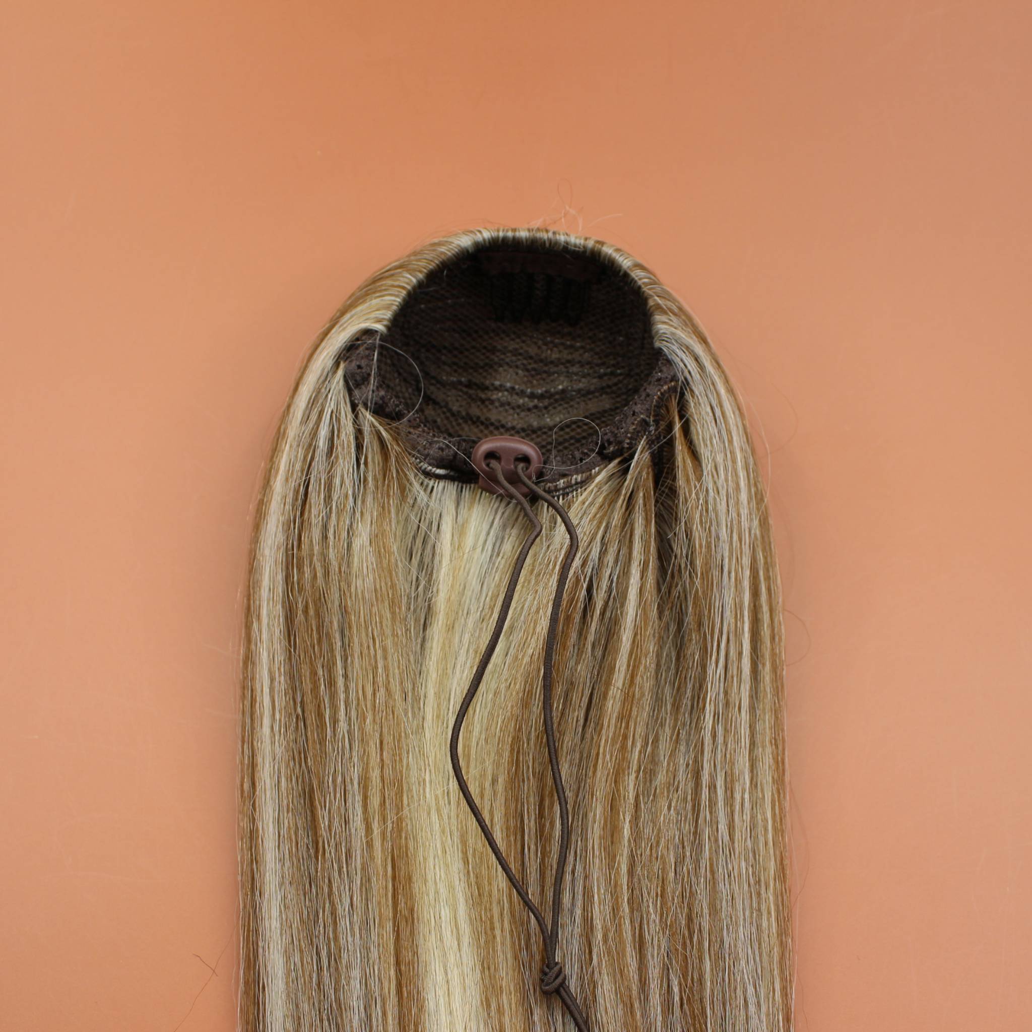DREAM HAIR Ponytail Straight 30'', Farbe 18/22 (blond mix) in der Rückansicht vor einem braun-orangen Hintergrund.