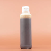 Das Hautpflegeprodukt SHEA GOLD Liquid Black Soap 240ml in der Rückansicht vor einem braun-orangen Hintergrund.