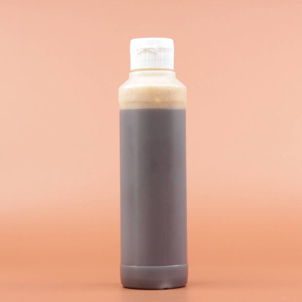 Das Hautpflegeprodukt SHEA GOLD Liquid Black Soap 240ml in der Rückansicht vor einem braun-orangen Hintergrund.