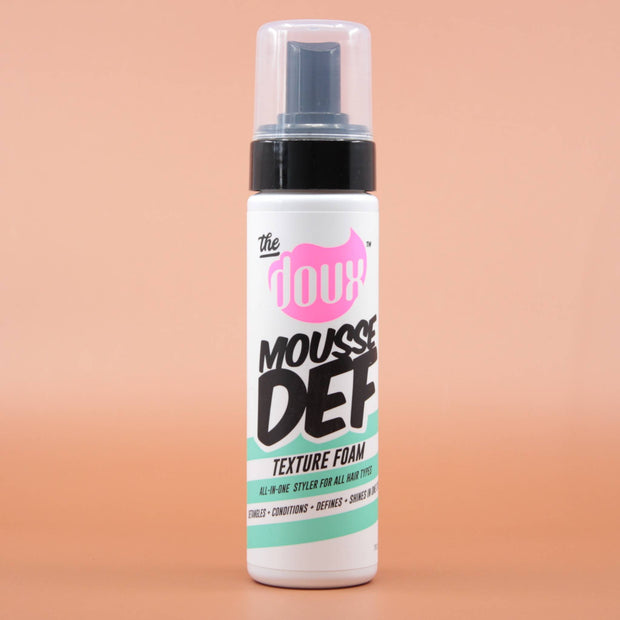 Das Haarstylingprodukt THE DOUX Mousse Def Texture Foam 207ml in der Vorderansicht vor einem braun-orangen Hintergrund.