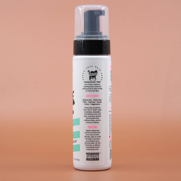 Das Haarstylingprodukt THE DOUX Mousse Def Texture Foam 207ml in der Seitenansicht (links) vor einem braun-orangen Hintergrund.